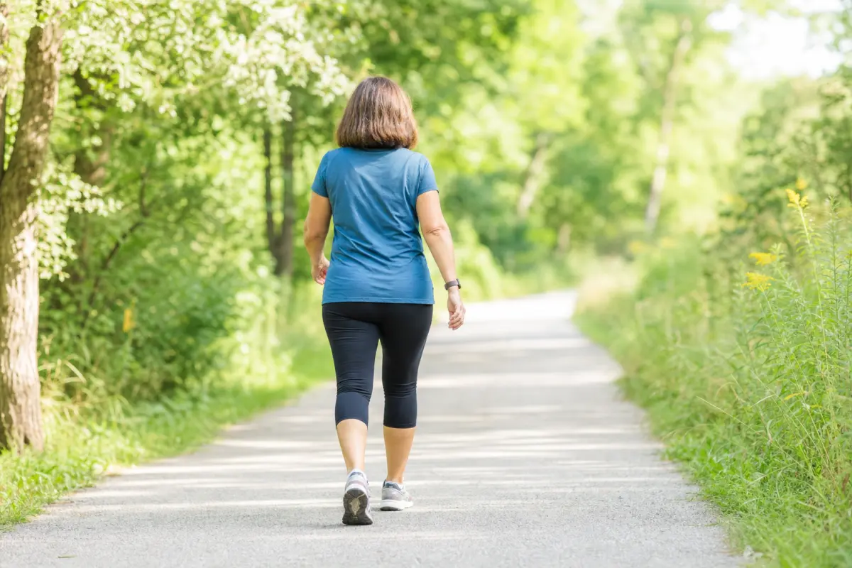 A woman walking briskly along a bright sunlit tree-lined path, photographed from behind